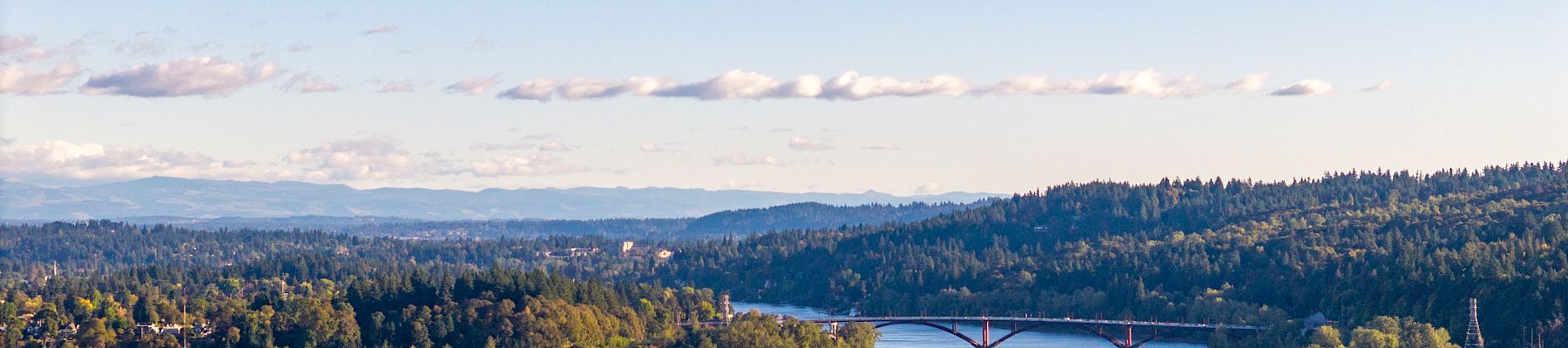 A wide river flows past forested hills, with sailboats, a marina, and a bridge stretching across under a blue sky.