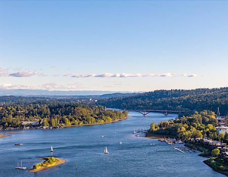 A wide river flows past forested hills, with sailboats, a marina, and a bridge stretching across under a blue sky.