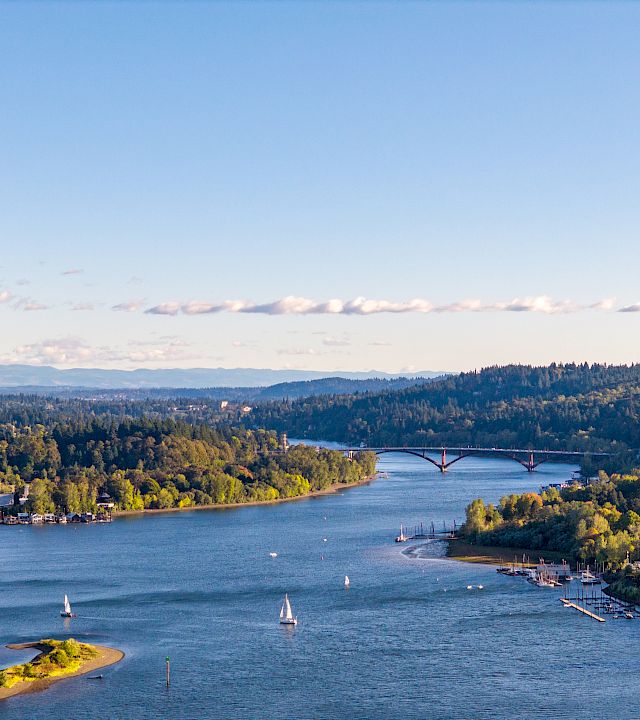 A wide river flows past forested hills, with sailboats, a marina, and a bridge stretching across under a blue sky.