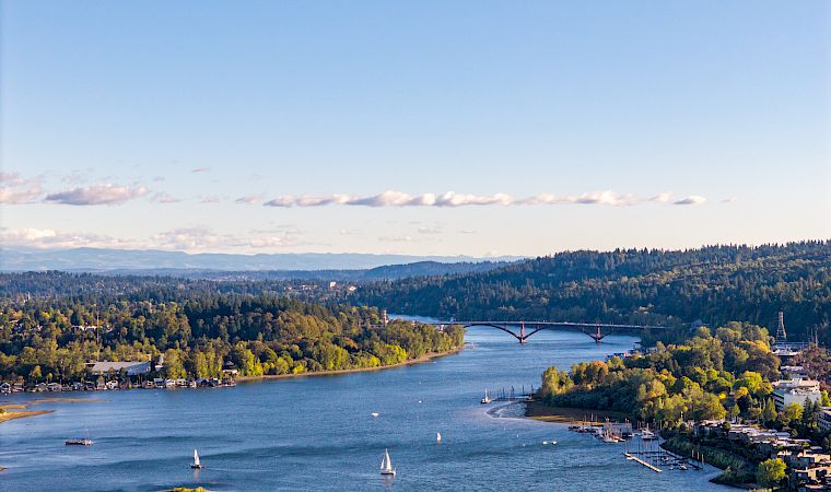 A wide river flows past forested hills, with sailboats, a marina, and a bridge stretching across under a blue sky.