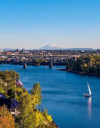 River at sunset with a steel bridge, sailboat, trees, and a distant snow-capped mountain under a blue sky.