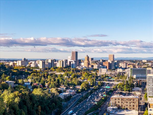 A sunny city skyline with tall office towers, busy highways, lots of trees, and a wide cloudy blue sky.